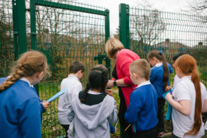 A teacher and a group of children are looking closely at a patch of grass behind a green metal mesh fence. The children are all holding light blue workbooks.