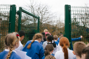 A group of school children are walking through a green metal mesh gate led by their schoolteacher.