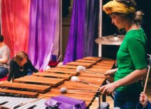 Plink and Boo production picture. A woman in a green top and yellow headband is playing a very large xylophone