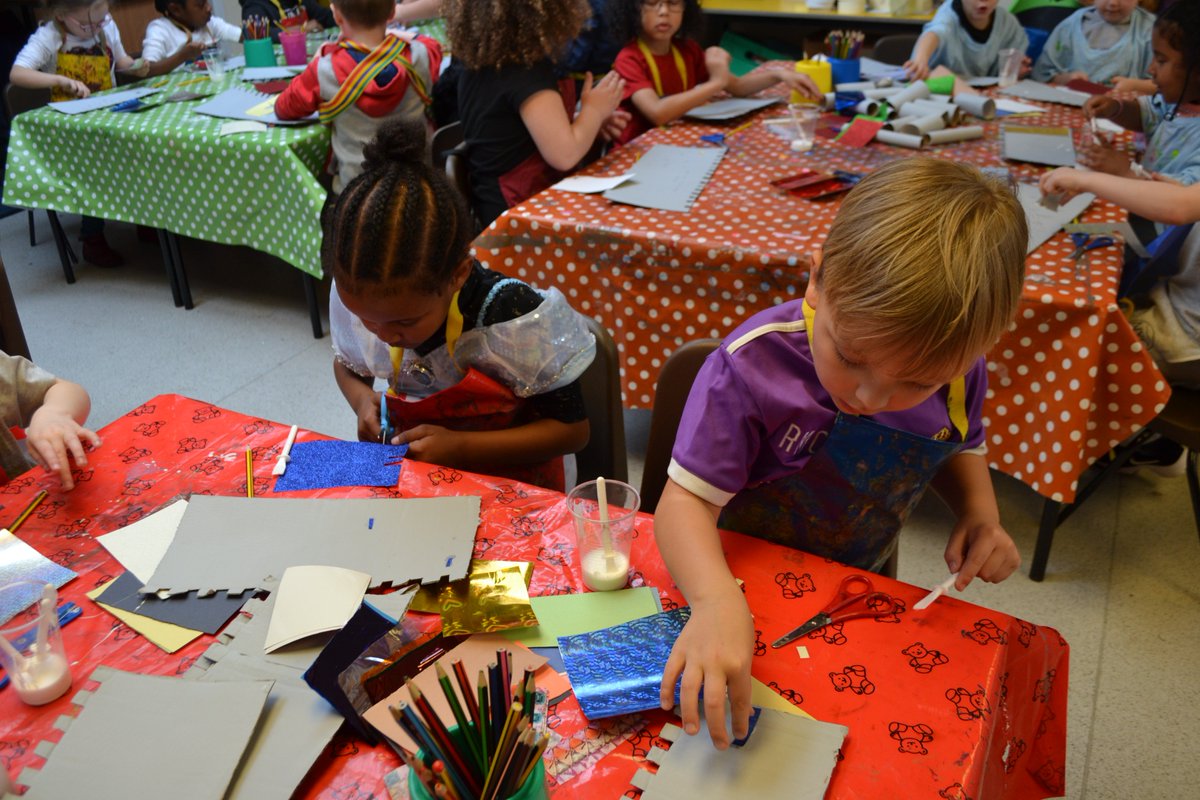 A boy and girl are doing art activities on a table with a red shiny, teddy bear-patterned table cloth. In the background there are two other groups of children doing the same activity on a red polkadot covered table and a green polkadot covered table