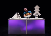 Production photo from 'A Square World'. A man at a light-up desk with lots of building blocks and three little squares with faces on.