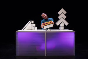 Production photo from 'A Square World'. A man at a light-up desk with lots of building blocks and three little squares with faces on.