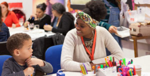 Z-arts Engagement Officer Yemi is wearing a cream cardigan and a colourful head band. She is sitting at a table and smiling at a young boy wearing a grey long-sleeved shirt sitting next to her. There are felt tip pens and scissors on the table.