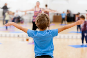 A young boy in a blue tshirt stands with his arms outstretched with his back to the camera