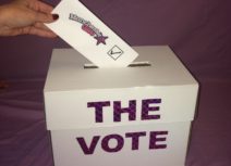 White Ballot box saying 'The Vote' in purple letters on the front with someone inserting a vote which reads 'Manchester Glee'