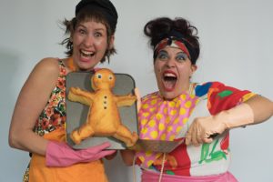 Two women smiling wearing colourful, mismatched clothing and holding a large gingerbread man style cake