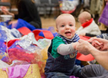 Photo of smiling baby holding an adult's hands. He is wearing a green and grey long-sleeved tshirt and blue denim dungarees. He is sitting in a pile of mixed texture, colourful fabric