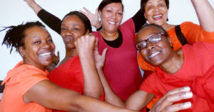 Five women wearing red tops. They are all looking at the camera, smiling and waving their arms