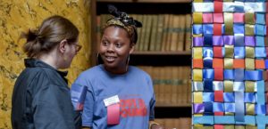 Photo of two women talking. The women on the left is wearing a navy and red 'Ministry of Lost and Found T-shirt' and is standing next to a lattice of different coloured ribbons.