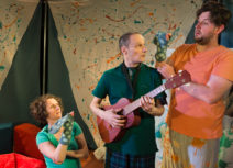 Production image from '5 More Minutes'. On the left, a lady in a green tshirt is kneeling on the floor with a green, spotty snake hand-puppet on her hand, next to her is a man in a green polo shirt holding a guitar, he is looking at a man next to him who also has a green, spotty snake hand-puppet and is wearing an orange t-shirt