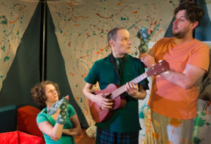 Production image from '5 More Minutes'. On the left, a lady in a green tshirt is kneeling on the floor with a green, spotty snake hand-puppet on her hand, next to her is a man in a green polo shirt holding a guitar, he is looking at a man next to him who also has a green, spotty snake hand-puppet and is wearing an orange t-shirt