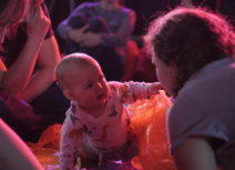 A baby wearing a long-sleeved animal print t-shirt is crawling through some orange mesh and pointing towards an adult wearing a grey tshirt