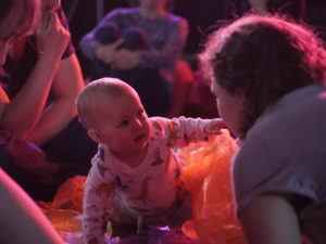 A baby wearing a long-sleeved animal print t-shirt is crawling through some orange mesh and pointing towards an adult wearing a grey tshirt