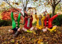 Production image for '4 Go Wild in Wellies'. Four adults are sitting cross-legged in a wooded area on a bed of Autumn leaves, they are all throwing leaves up into the air and are wearing scarves, wellies and colourful raincoats