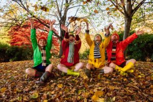 Production image for '4 Go Wild in Wellies'. Four adults are sitting cross-legged in a wooded area on a bed of Autumn leaves, they are all throwing leaves up into the air and are wearing scarves, wellies and colourful raincoats