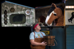 Production image from 'Black Beauty' showing a man holding a bucket and wearing a blue t-shirt looking up at a brown horse with a white nose who is standing inside a horse box.