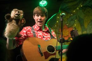 Production image from 'Climb That Tree' - David Gibb, a man in a red shirt is holding a guitar and a brown hand puppet of a small animal.