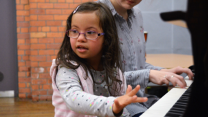 A girl approximately 6 years old is sitting at a piano. She is wearing a grey polkadot jumper and a light pink stripey gilet. Next to her is an adult female wearing a black and white stripey shirt