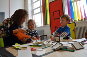 An adult and a boy and girl aged approximately 4 years are sitting doing art activities at a table. Felt tips, scissors, tin foil and paper is on the table.