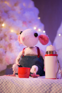 Production image from 'Snow Mouse' of a white toy mouse with pink ears sitting next to a white flask. The mouse has pink feet and is sitting in front of a red cup.