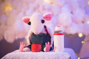 Production image from 'Snow Mouse' of a white toy mouse with pink ears sitting next to a white flask. The mouse has pink feet and is sitting in front of a red cup.