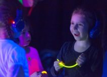 Three children wearing headphones and holding glowsticks. Their clothes are glowing as they are standing under UV light. You can see one child's face clearly, she is blonde and has a happy and surprised expression on her face.