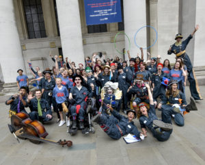 Photo of all the cast and volunteers of 'The Ministry of Lost and Found', a lot of them are waving at the camera and they are outside Manchester Central Library and they are all wearing navy blue. There are approximately 50 people in the photo and there is a double bass lying on the floor in front of the people on the left hand side