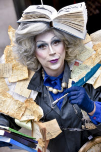 An adult is dressed in character with white facepaint, purple lipstick and blue eyeshadow and a costume made of books. The character was part of The Ministry of Lost and Found and Manchester Central Library.