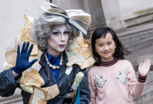 A girl in a pink jumper with black hair is standing next to an adult character with white facepaint and a costume made of books.