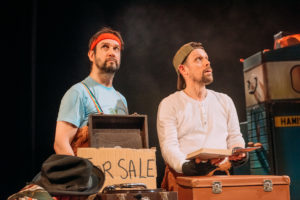 Production image from Black Beauty of two middle aged men with brown hair. One is wearing a blue tshirt and a red headband, one is wearing a white long-sleeved tshirt and a khaki baseball cap worn backwards.