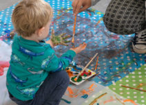 A toddler wearing a green, dinosaur-print tshirt is crouched on plastic table cloths which are covered in paint. An adult is crouching in front of him painting more paint onto his hands with a paintbrush.