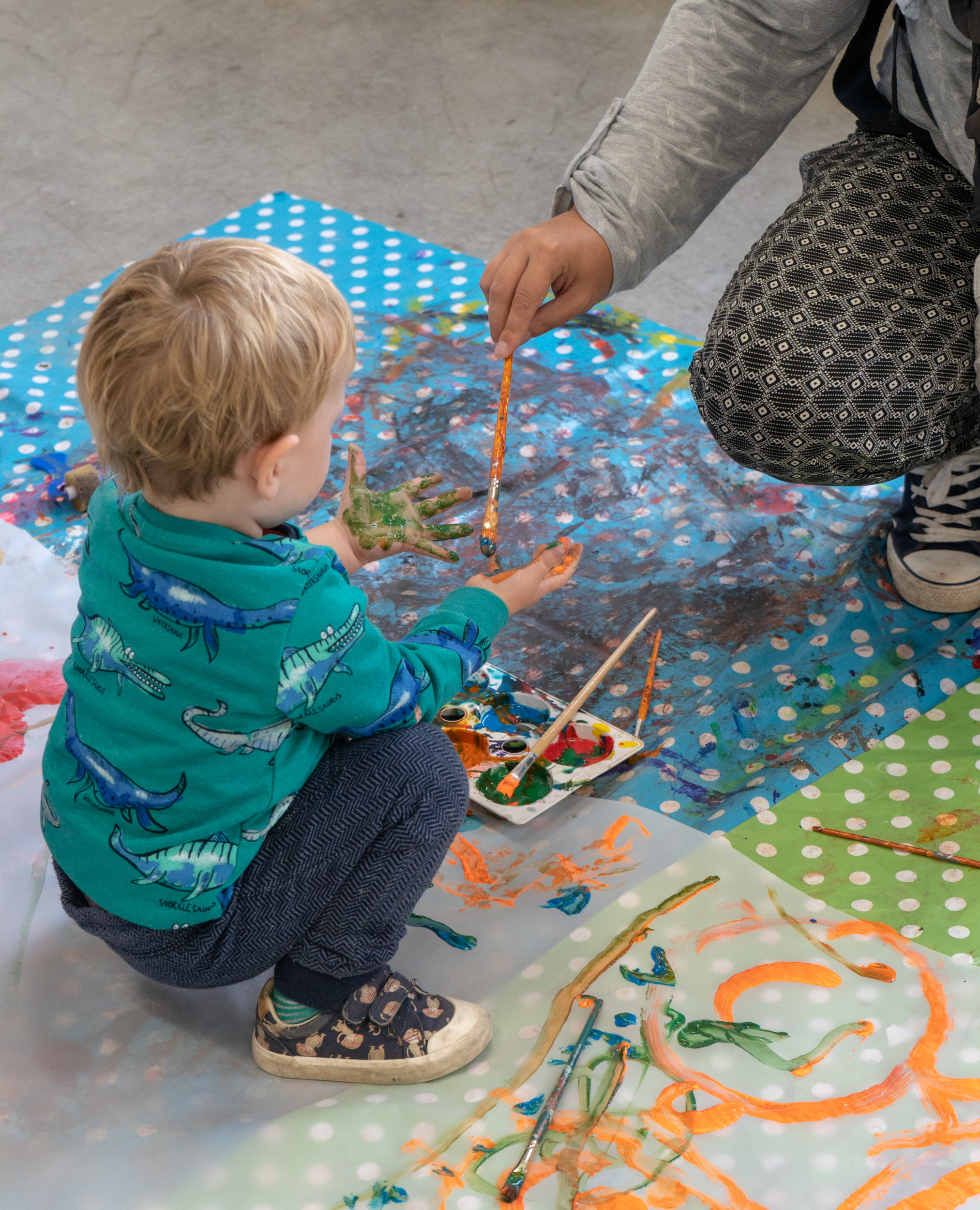 A toddler wearing a green, dinosaur-print tshirt is crouched on plastic table cloths which are covered in paint. An adult is crouching in front of him painting more paint onto his hands with a paintbrush.