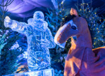 Picture of pantomime horse who is brown with a white nose and white lower legs, the horse is standing in front of a sculpture made of ice and a Christmas tree