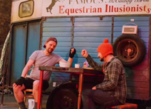 Production image from 'Black Beauty', they are both sitting down in front of a blue horse box and are bumping fists. One man is wearing a red bobble hat and a checked shirt, one is wearing a black baseball cap worn backwards and a grey tshirt