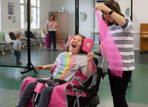 Image of woman who is a wheelchair user laughing with a woman standing next to her with a piece of fabric