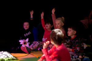 Photo from a previous Drag Queen Story Time show. Photoof five young children sitting on the floor with their hands raised in the air