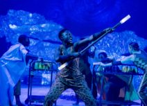 Production photo from The Little Prince, the stage is lit in blue - in the foreground a women in a printed jumpsuit is brandishing a large stick with illuminated ends. Her facial expression looks as though she is shouting.