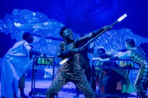 Production photo from The Little Prince, the stage is lit in blue - in the foreground a women in a printed jumpsuit is brandishing a large stick with illuminated ends. Her facial expression looks as though she is shouting.