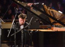 Photo of a man sat playing a grand piano. He has short brown hair and is wearing a tuxedo.