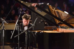 Photo of a man sat playing a grand piano. He has short brown hair and is wearing a tuxedo.