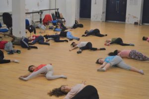 Picture of a group of teenagers lying on the floor of a dance studio in a yoga stretch pushing one leg over to the side