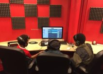 Photo of three boys sat in front of a mac desktop computer. There is a red and black check patterned wall in the background