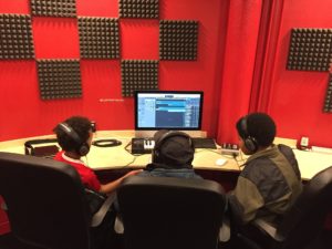 Photo of three boys sat in front of a mac desktop computer. There is a red and black check patterned wall in the background
