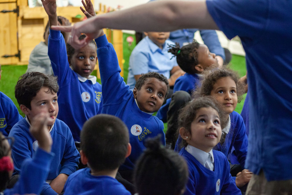 Photo of schoolchildren sitting on the floor. They are wearing school uniform of a blue jumper and blue shirts underneath.