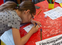 Girl with long brown hair and a grey spotty tshirt leaning over a table drawing on a piece of paper