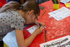 Girl with long brown hair and a grey spotty tshirt leaning over a table drawing on a piece of paper