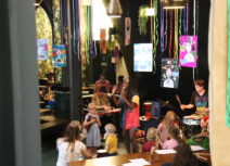 One Manchester Family Jam promo image. A woman is standing singing in front of an audience of young families. She is wearing a colourful dress and there are multicoloured streamers floating down from the ceiling above her head.