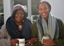 Picture of two women attending Community Coffee Morning. They are both holding mugs of coffee, they are both wearing wool scarves and smiling.