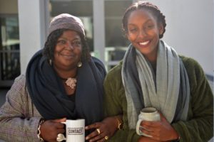 Picture of two women attending Community Coffee Morning. They are both holding mugs of coffee, they are both wearing wool scarves and smiling.