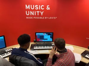 Lead image for songwriting competition. Two young people are sitting in front of a music keyboard and a computer screen, behind the screen is a red wall that says "music and unity" on it.